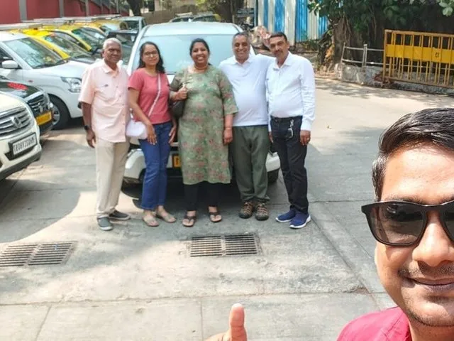 A group of tourists smiling with their driver next to a white car.